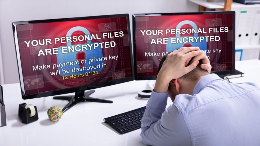 A person sits at a desk holding their head, facing two monitors displaying a ransomware message stating, "Your personal files are encrypted," highlighting the dangers of ransomware spread across networks via WiFi.