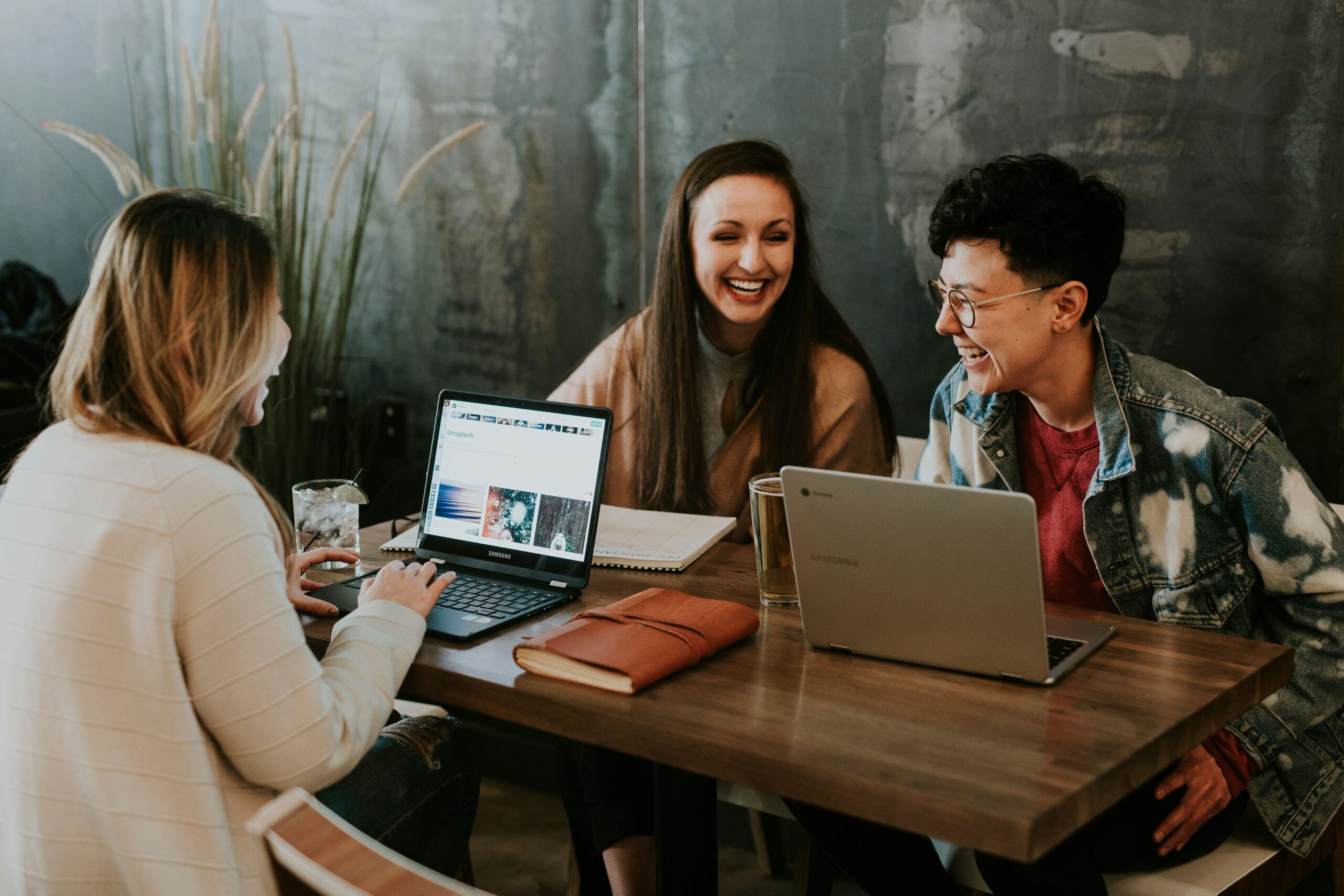 Three people sitting at a wooden table, discussing and laughing, with laptops and notebooks in front of them for their meeting about purchasing Office 365 subscription.